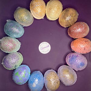 May include: A circular arrangement of fourteen oval bath bombs with a textured, scale-like surface in various colors including blue, green, purple, orange, and gold. A small white circular label in the center reads Aura Apothecary.