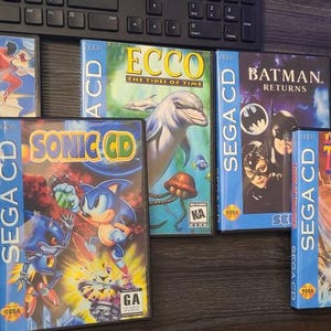 May include: A collection of SEGA CD video game cases, including "Final Fight," "Sonic CD," "Ecco the Tides of Time," "Batman Returns," "Lords of Thunder," and "The Terminator." The cases are blue with game titles and artwork.