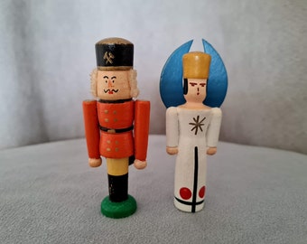 May include: A wooden nutcracker figurine wearing a black hat with a gold hammer and pick symbol, a red jacket, yellow trousers, and black boots on a green base, alongside a wooden angel figurine with a gold crown, blue wings, and white robe.