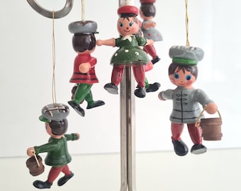 May include: Set of six wooden ornaments depicting people wearing hats and carrying buckets. The ornaments are painted in various colors, including red, green, blue, and gray. The ornaments are hanging from a metal stand.