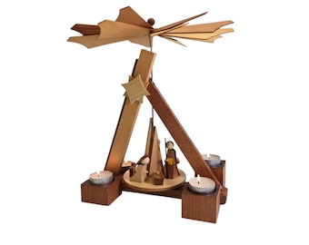 May include: A wooden Christmas pyramid featuring a natural light wood finish and darker wood accents. The structure includes a rotating top fan, a central nativity scene with stylized figures, and three candle holders with lit tea light candles.