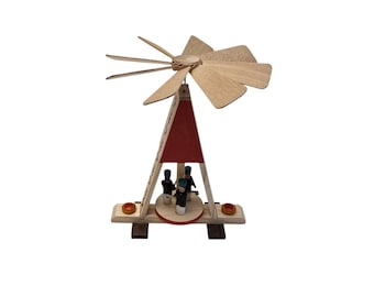 May include: A wooden Christmas pyramid featuring a natural wood frame with a red triangular center section. At the base, three small figures in dark uniforms stand on a rotating platform. A large wooden propeller sits at the top of the structure.