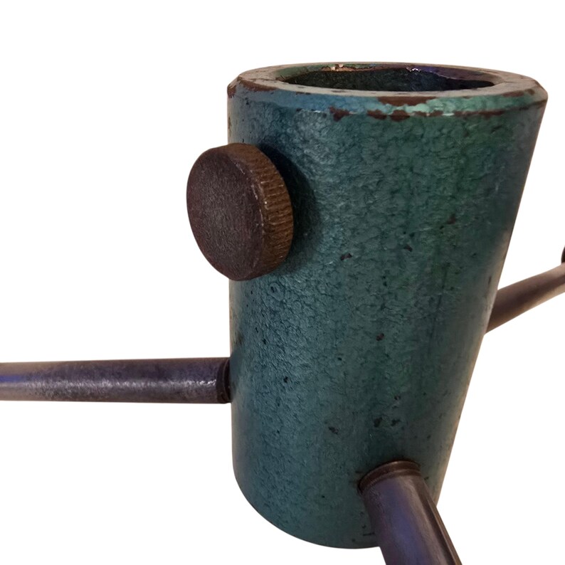 May include: A teal-colored metal Christmas tree stand with a textured finish. The stand has a round, dark brown knob and three metal legs extending outward. The stand is designed to hold a Christmas tree.