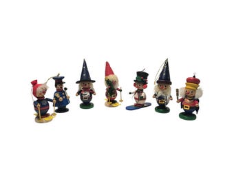 May include: A collection of seven small wooden ornaments featuring various characters including skiers, a wizard, a snowman, and a soldier. Each figure is painted in vibrant colors and includes a small string loop for hanging as a decoration.