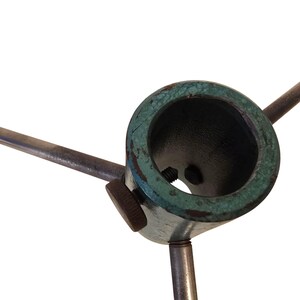 May include: Close-up of a metal tripod base with a circular top and three supporting legs. The top has a dark interior and a weathered, teal-colored rim. A small, round knob is visible on the side. The metal has a worn, aged appearance.