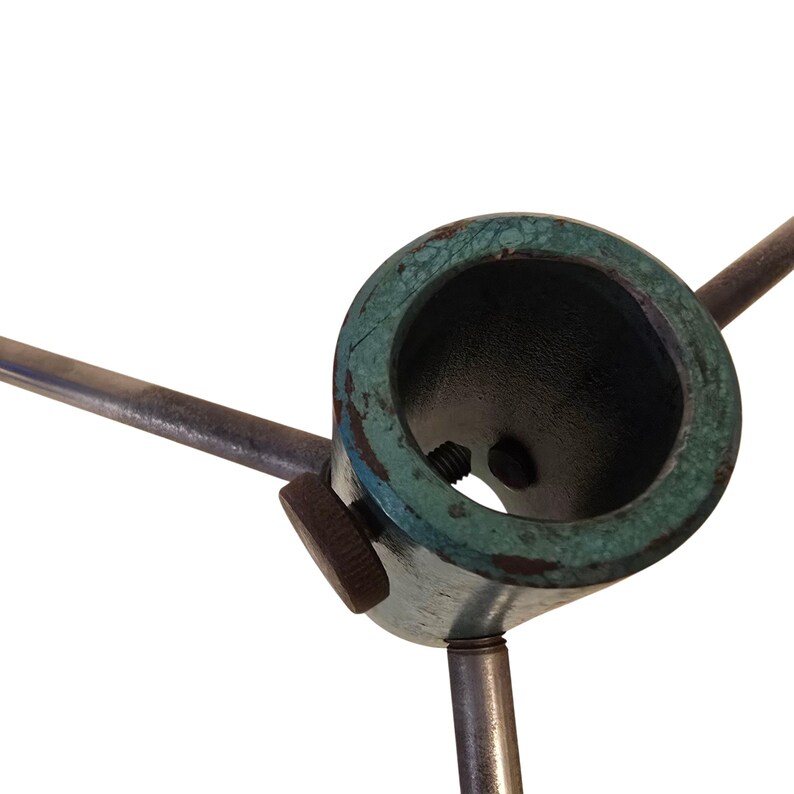 May include: Close-up of a metal tripod base with a circular top and three supporting legs. The top has a dark interior and a weathered, teal-colored rim. A small, round knob is visible on the side. The metal has a worn, aged appearance.