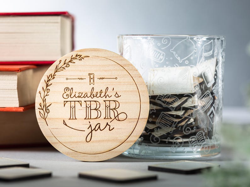 May include: A wooden round coaster with the text "Elizabeth's TBR jar" engraved on it. The coaster is sitting on top of a glass jar with a design of books and other reading-related items.