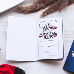 May include: A white passport-style booklet with a red and black logo that says "Monorail Crawl 2022". The logo features a cartoon image of a monorail. The booklet has a section for passenger information with the words "Name" and "Date" printed on it.