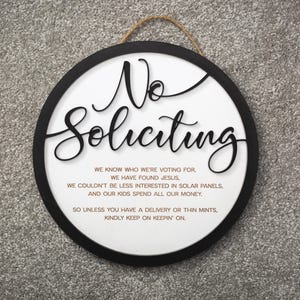 May include: A round, black-framed sign with white background and the text "No Soliciting" in large, black, cursive lettering. The sign also includes a smaller text message in black lettering that reads: "We know who we're voting for, we have found Jesus, we couldn't be less interested in solar panels, and our kids spend all our money. So unless you have a delivery or thin mints, kindly keep on keepin' on."