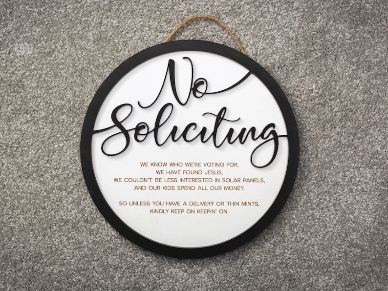 May include: A round, black-framed sign with white background and the text "No Soliciting" in large, black, cursive lettering. The sign also includes a smaller text message in black lettering that reads: "We know who we're voting for, we have found Jesus, we couldn't be less interested in solar panels, and our kids spend all our money. So unless you have a delivery or thin mints, kindly keep on keepin' on."