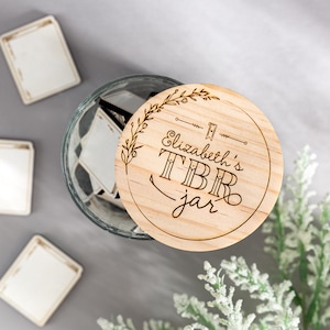 May include: A wooden jar lid with the text "Elizabeth's TBR jar" engraved on it. The lid is decorated with a floral design and a small flag.