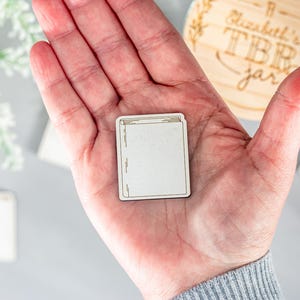 May include: A wooden book-shaped token with a blank white surface. The token is being held in a person's hand.