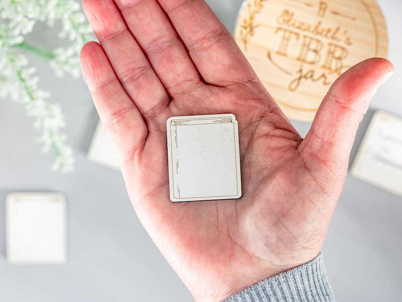 May include: A wooden book-shaped token with a blank white surface. The token is being held in a person's hand.