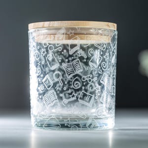 May include: A clear glass jar with a wooden lid. The jar is decorated with a white etched design of books, stars, and other school-related items.