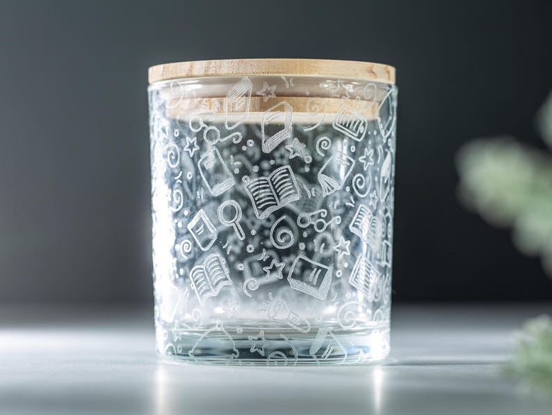 May include: A clear glass jar with a wooden lid. The jar is decorated with a white etched design of books, stars, and other school-related items.