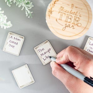 May include: A wooden jar with the text "Elizabeth's TBR jar" written on it.  There are several small wooden cards with book titles written on them, including "A Man Called Ove", "The Midnight Lib", and "Lessons in Chemistry".