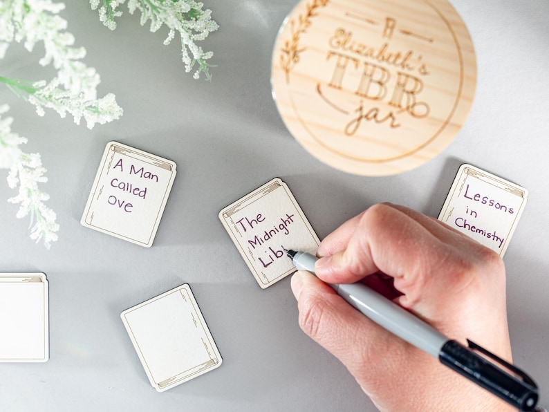 May include: A wooden jar with the text "Elizabeth's TBR jar" written on it.  There are several small wooden cards with book titles written on them, including "A Man Called Ove", "The Midnight Lib", and "Lessons in Chemistry".