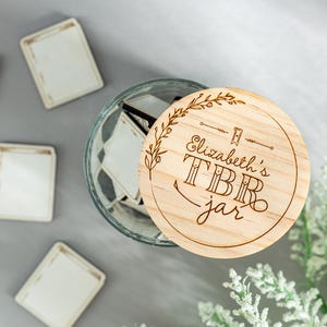 May include: A wooden jar lid with the text "Elizabeth's TBR jar" engraved on it. The lid is decorated with a floral design and is sitting on top of a glass jar filled with small white cards.
