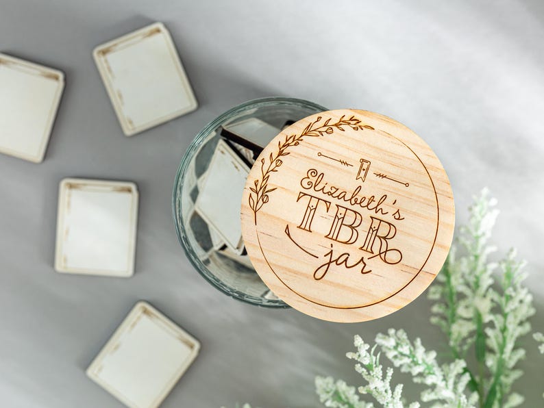 May include: A wooden jar lid with the text "Elizabeth's TBR jar" engraved on it. The lid is decorated with a floral design and is sitting on top of a glass jar filled with small white cards.