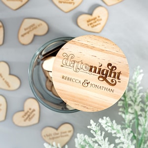 May include: A glass jar filled with wooden heart-shaped date night ideas. The lid is made of wood and has the text "datenight" with an arrow and a crescent moon. The names "Rebecca & Jonathan" are also engraved on the lid.