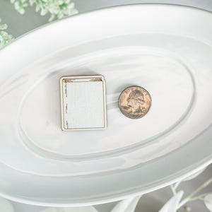 May include: A white ceramic oval serving dish with a wooden book-shaped ornament and a silver quarter on a white surface.