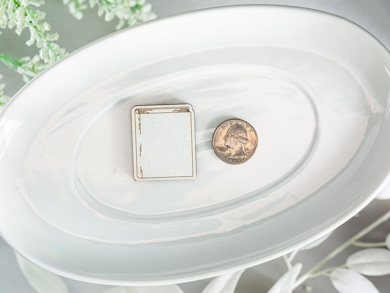 May include: A white ceramic oval serving dish with a wooden book-shaped ornament and a silver quarter on a white surface.