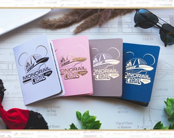 May include: Four small, square notebooks with a gold foil design of a monorail and the text "Monorail Crawl 2022". The notebooks are in white, pink, grey, and blue.