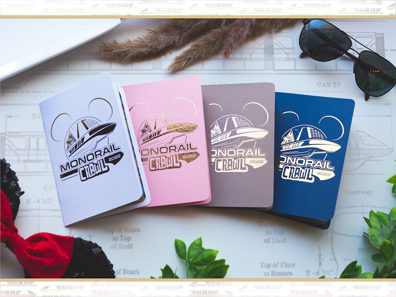 May include: Four small, square notebooks with a gold foil design of a monorail and the text "Monorail Crawl 2022". The notebooks are in white, pink, grey, and blue.