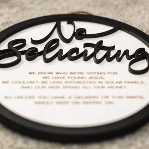 May include: A black and white round sign with the text "No Soliciting" in a decorative font. The sign also includes a short message about voting, finding Jesus, and solar panels.