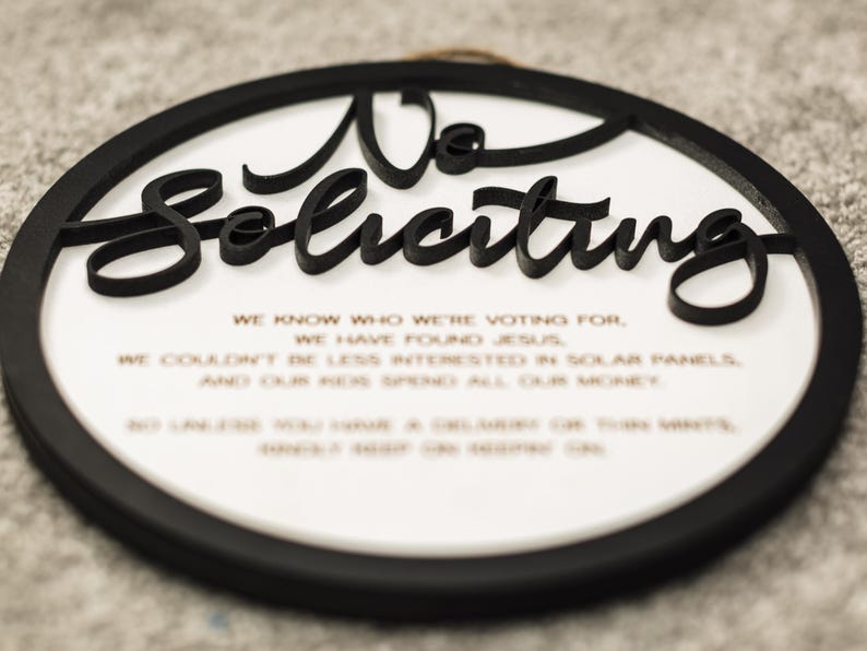 May include: A black and white round sign with the text "No Soliciting" in a decorative font. The sign also includes a short message about voting, finding Jesus, and solar panels.