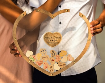 Könnte beinhalten: Ein herzförmiger Rahmen aus Holz mit einem klaren Acryleinsatz. Der Rahmen ist mit goldfarbenen Tackern verziert und hat eine Holzherzplakette im Inneren mit dem Text "James & Charlotte September 4, 2021". Der Rahmen ist mit kleinen Holzh Herzen gefüllt, auf denen handgeschriebene Botschaften stehen.