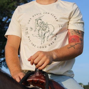 May include: A person wearing a cream colored t-shirt with a green graphic of a cowboy riding a bucking bronco. The graphic is surrounded by text that reads "The Devil Can Scrap But The Lord Has Won 2023".