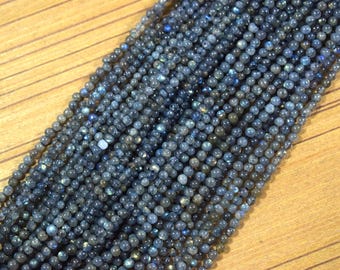 May include: Close-up of strands of small, round, blue-grey beads. The beads have a subtle iridescence and are strung together, creating a textured surface. The background is a light brown wooden surface.