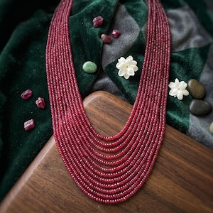 May include: A multi-strand necklace composed of numerous small, faceted, deep red gemstone beads arranged in a cascading, layered design. The necklace features a consistent, uniform texture created by the repetitive pattern of the circular beads.