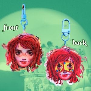 Puede incluir: Un llavero de doble cara con una mujer de dibujos animados de pelo rojo. La parte delantera muestra la cara de la mujer, mientras que la parte trasera la muestra con una máscara amarilla con detalles rojos y azules.