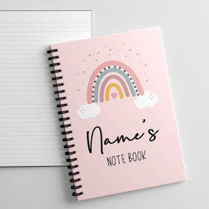 May include: Pink notebook featuring a rainbow, clouds, and stars design. The notebook has a spiral binding and the text "Name's NOTE BOOK" on the front.