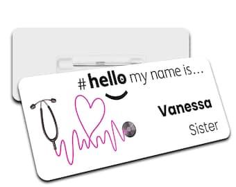May include: White name tag with a black hashtag, the text "#hello my name is...", a pink heart, a stethoscope, a pink EKG line, a black record, the name "Vanessa" and the word "Sister".