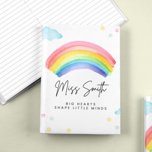 May include: A white notebook featuring a watercolour rainbow design. The cover displays the name "Miss Smith" in black script, with the text "Big Hearts Shape Little Minds" below. Pastel clouds and dots decorate the cover.