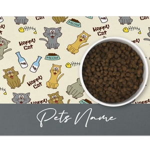 May include: A beige and gray cat-themed pet bowl mat with a white bowl filled with brown kibble. The mat has a repeating pattern of cats, fish bones, milk bottles, and the words "Happy Cat" and "Meow". The bottom of the mat is gray and has the text "Pets Name" written in white.