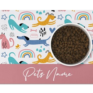 May include: White dog bowl with brown kibble on a pink and white dog bowl mat with a rainbow, stars, and dog illustrations. The mat has the text "Pets Name" in pink.