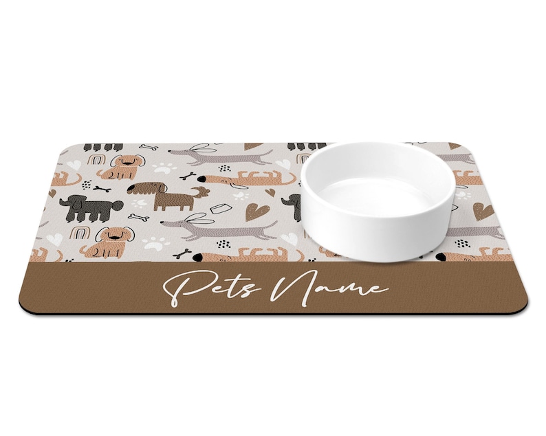 May include: A brown and white dog-themed pet bowl mat with a white ceramic bowl. The mat has a brown border with the text "Pets Name" in white.