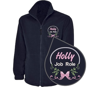 May include: Dark navy fleece jacket with a full zip and a high collar. The jacket has an embroidered design on the chest with the name "Holly" and "Job Role" in pink, framed by a pearl border and floral details.