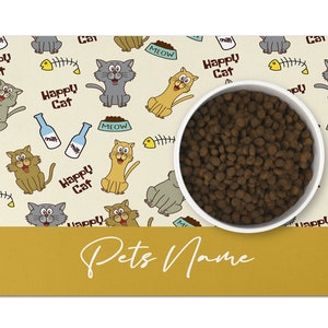 May include: A beige and yellow cat-themed pet bowl mat with a white bowl filled with brown kibble. The mat has a repeating pattern of cats, fish bones, milk bottles, and bowls that say "MEOW". The text "Happy Cat" is repeated throughout the mat. The bottom of the mat has a yellow bar with the text "Pets Name" in white.