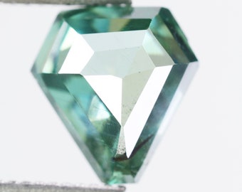 May include: A shield-shaped loose gemstone with a faceted cut, displaying a translucent teal-green color. The stone is held by a pair of metal tweezers, showcasing its geometric shape and reflective surfaces against a plain white background.