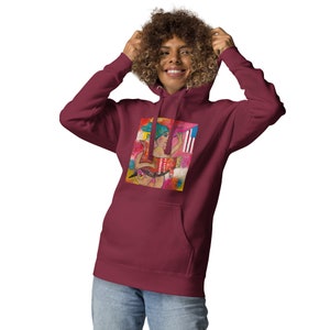 May include: A maroon hoodie with a colorful graphic print featuring a woman holding a paintbrush and a flag. The graphic is set against a pink and yellow background.