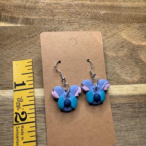 May include: A pair of handmade earrings featuring a blue and purple character design with silver-colored ear wires. The earrings are displayed on a brown card with a measuring tape in the background.