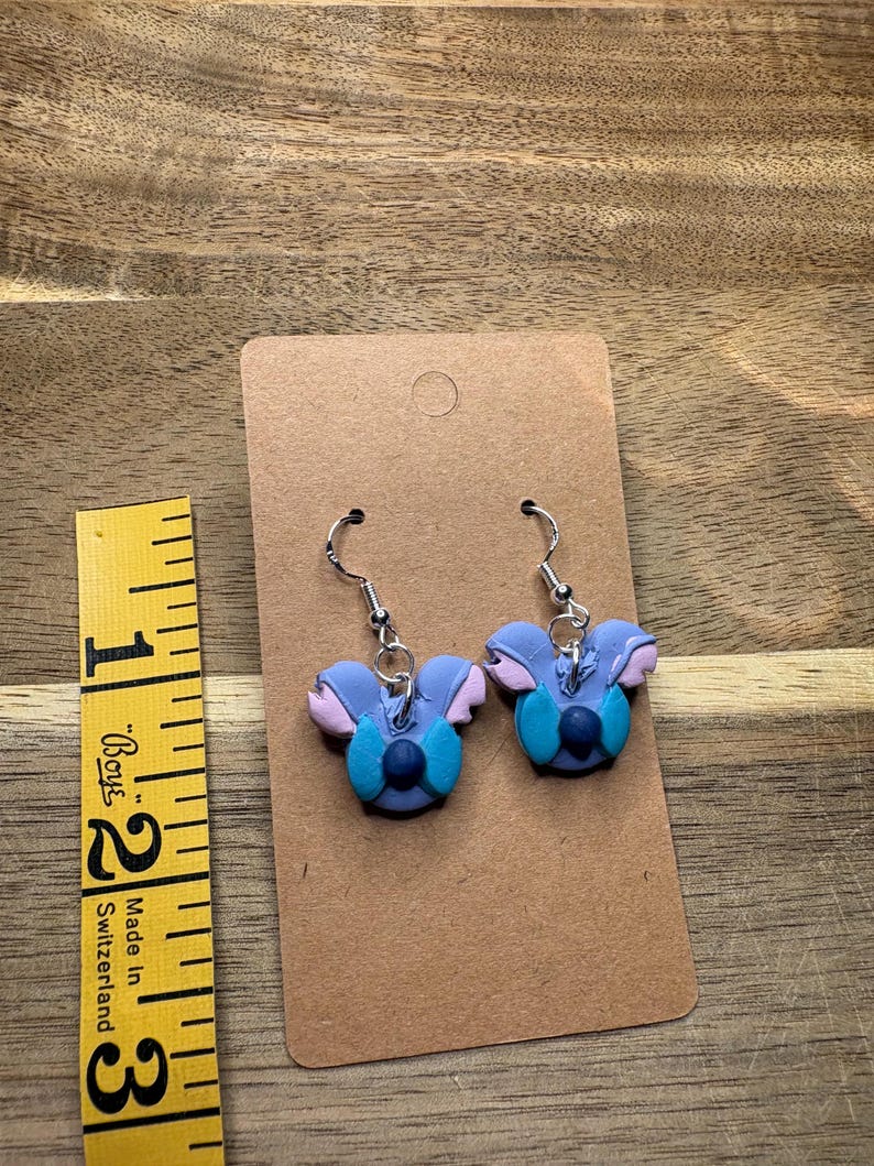 May include: A pair of handmade earrings featuring a blue and purple character design with silver-colored ear wires. The earrings are displayed on a brown card with a measuring tape in the background.