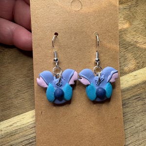 May include: A pair of handmade earrings featuring a blue, pink, and teal floral design. The earrings are made of polymer clay and have silver-colored ear wires. The earrings are displayed on a brown card.