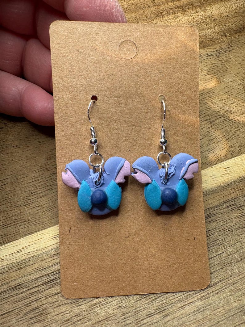 May include: A pair of handmade earrings featuring a blue, pink, and teal floral design. The earrings are made of polymer clay and have silver-colored ear wires. The earrings are displayed on a brown card.