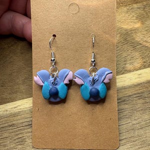 May include: A pair of handmade earrings featuring a blue, pink, and teal character design. The earrings are attached to silver-colored fish hook ear wires and are displayed on a brown card.
